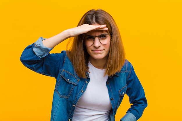 Jeune jolie femme à la perplexité et étonné, avec la main sur le front à la recherche au loin, regarder ou chercher contre le mur jaune