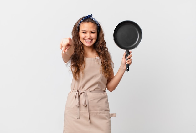 Jeune jolie femme pointant sur la caméra vous choisissant le concept de chef et tenant une casserole