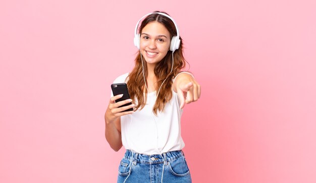 Jeune jolie femme pointant sur la caméra vous choisissant avec des écouteurs et un smartphone