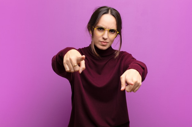 Jeune jolie femme pointant vers l'avant à la caméra avec les deux doigts et l'expression en colère, vous disant de faire votre devoir contre le mur violet