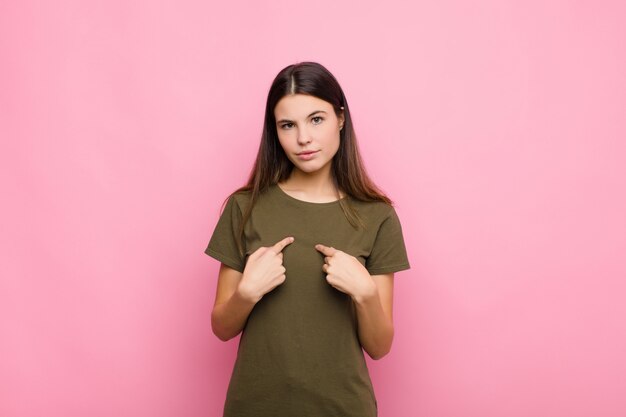 Jeune jolie femme pointant vers soi avec un regard confus et perplexe, choquée et surprise d'être choisie contre le mur rose