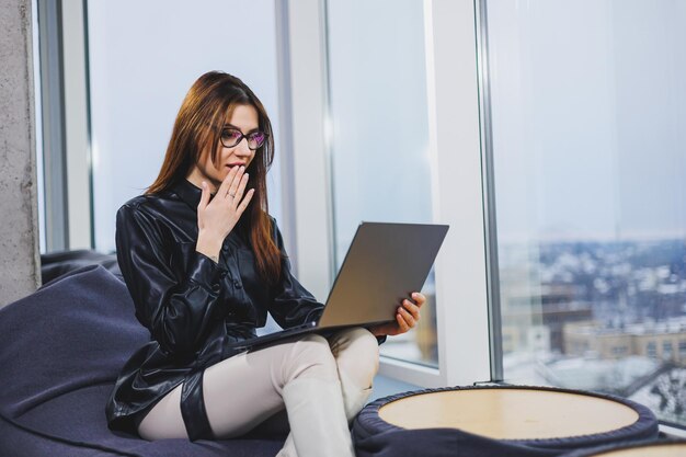 Une jeune jolie femme portant des lunettes travaille sur un ordinateur portable tout en étant assise dans un espace de travail moderne