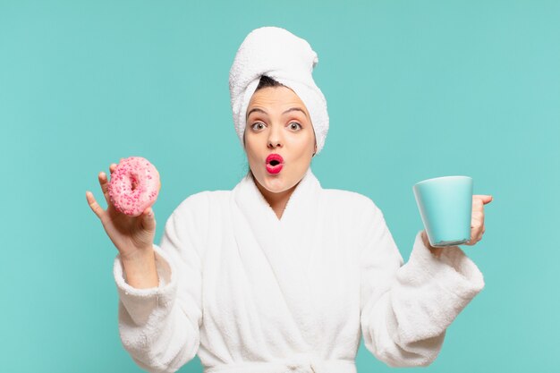 Jeune jolie femme portant un peignoir avec une expression surprise et prenant un petit-déjeuner