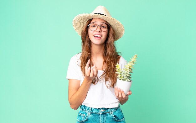 Jeune jolie femme à la recherche désespérée, frustrée et stressée avec un chapeau de paille et tenant un cactus