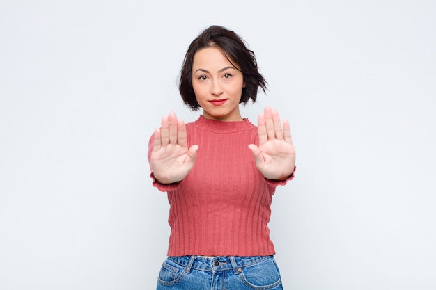 Jeune jolie femme à la recherche sérieuse, malheureuse, en colère et mécontente interdisant l'entrée ou disant arrêter avec les deux paumes ouvertes contre le mur blanc