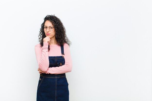 Jeune jolie femme à la recherche de sérieux, confus, incertain et réfléchi, doutant parmi les options ou les choix contre le mur blanc