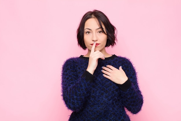 Jeune jolie femme à la recherche de sérieux et de croix avec le doigt pressé sur les lèvres exigeant le silence ou le calme, gardant un secret contre le mur rose