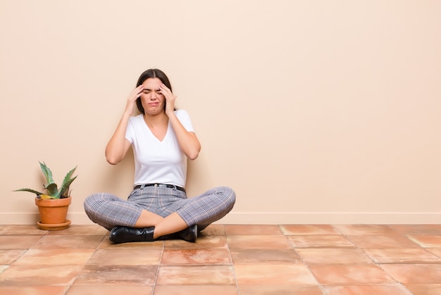 Jeune jolie femme à la recherche de stress et de frustration, travaillant sous pression avec un mal de tête et troublé par des problèmes assis sur un sol