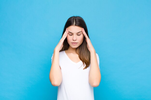 Jeune jolie femme à la recherche de stress et de frustration, travaillant sous pression avec un mal de tête et troublé par des problèmes sur le mur bleu