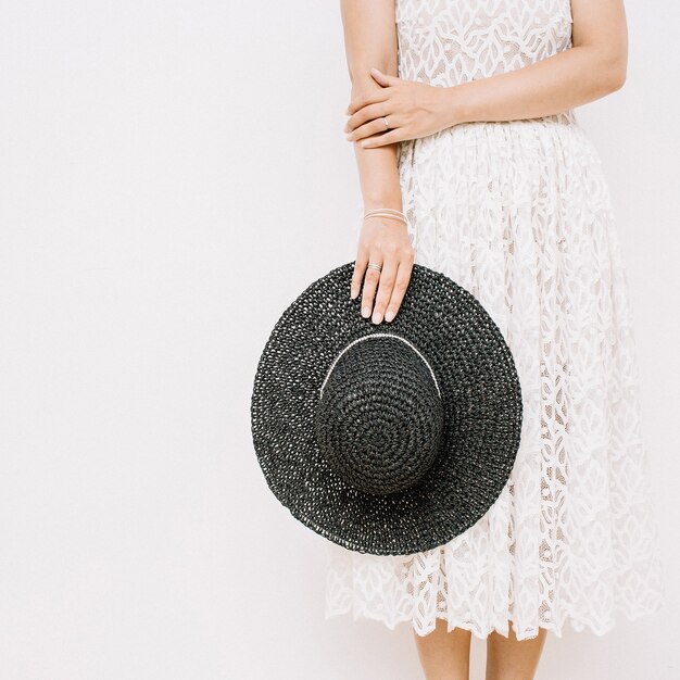 Jeune jolie femme en robe blanche avec un chapeau noir près du mur blanc