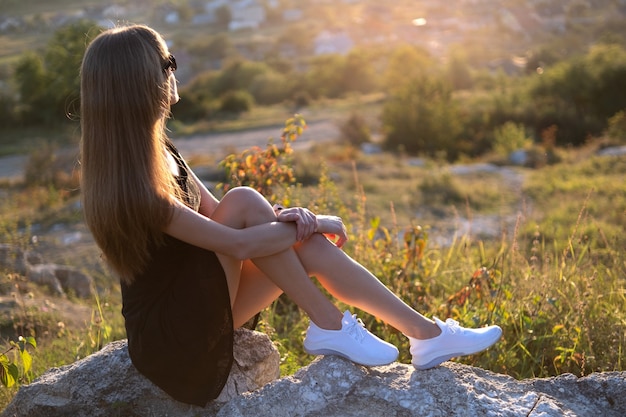 Jeune jolie femme en robe d'été courte noire assise sur un rocher relaxant à l'extérieur au coucher du soleil.