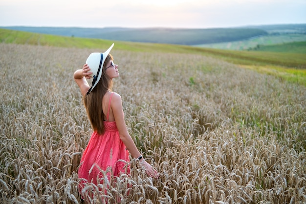 Jeune jolie femme en robe d'été rouge et chapeau de paille debout sur un champ de ferme jaune avec du blé doré mûr profitant d'une soirée chaude.