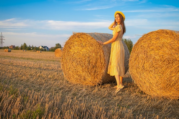 Jeune jolie femme en robe jaune et chapeau gros plan sur le fond du champ avec des meules de foin au coucher du soleil