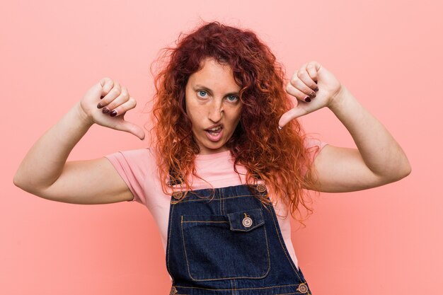 Jeune jolie femme rousse au gingembre portant une salopette en jean montrant le pouce vers le bas et exprimant l'aversion