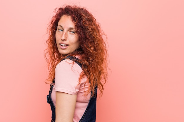 Jeune jolie femme rousse au gingembre portant une salopette en jean regarde de côté souriant, gai et agréable.