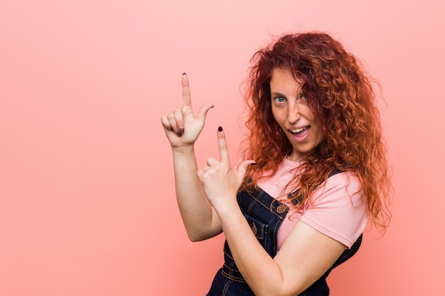 Jeune jolie femme rousse au gingembre portant une salopette en jeans pointant avec les index vers un espace de copie, exprimant l'excitation et le désir.