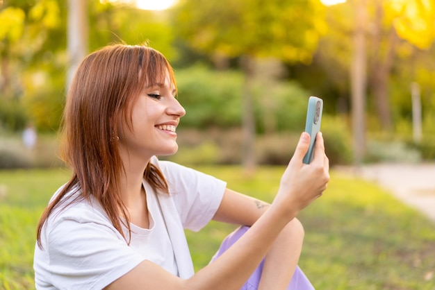 Jeune jolie femme rousse à l'extérieur à l'aide d'un téléphone portable avec une expression heureuse