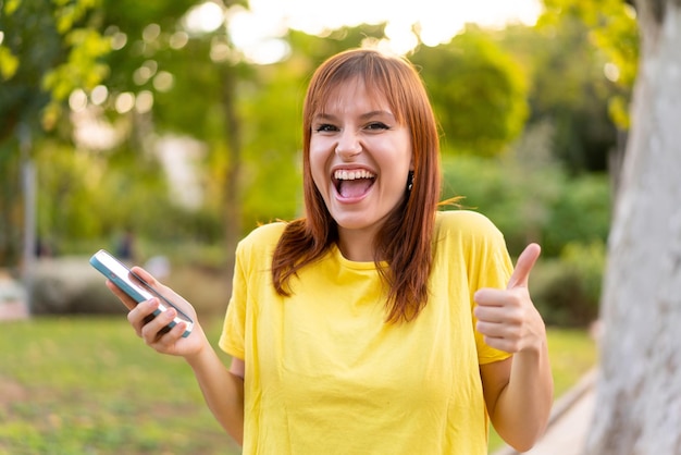 Jeune jolie femme rousse à l'extérieur à l'aide d'un téléphone portable tout en faisant des pouces vers le haut