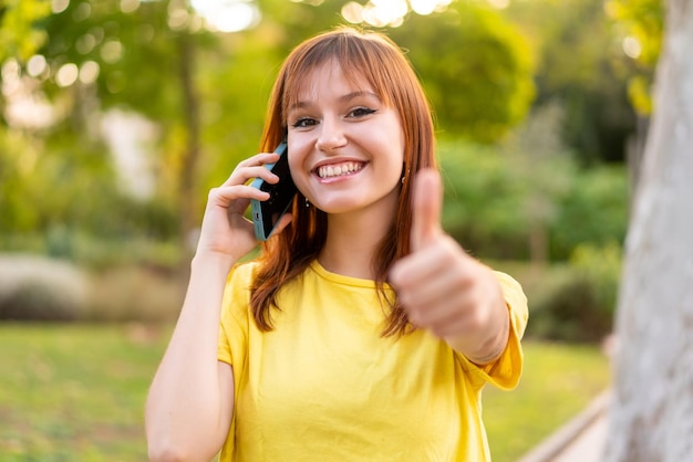 Jeune jolie femme rousse à l'extérieur en gardant une conversation avec le mobile tout en faisant des pouces vers le haut