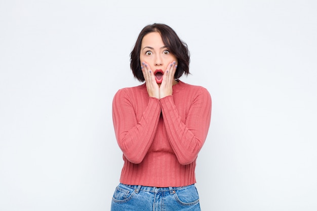 Jeune jolie femme se sentant choquée et effrayée, terrifiée avec la bouche ouverte et les mains sur les joues sur le mur blanc