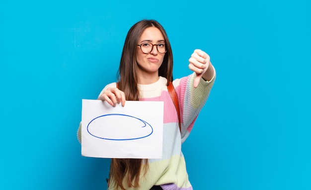 Jeune jolie femme se sentant fâchée, en colère, agacée, déçue ou mécontente, montrant les pouces vers le bas avec un regard sérieux