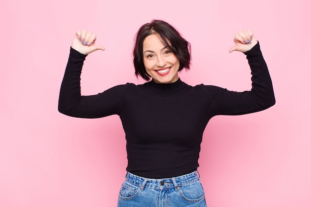 Jeune jolie femme se sentant fière, arrogante et confiante, semblant satisfaite et réussie, pointant vers soi contre le mur rose