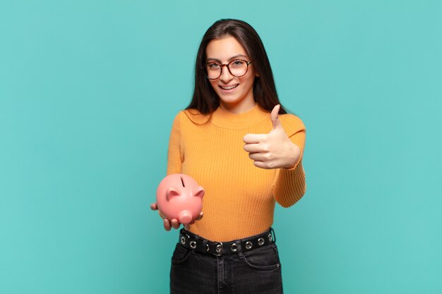 Jeune jolie femme se sentant fière, insouciante, confiante et heureuse, souriant positivement avec le pouce levé. concept de tirelire