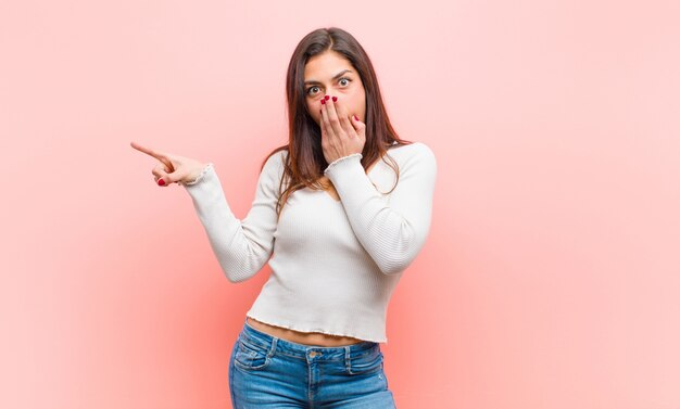 Jeune jolie femme se sentant heureuse, choquée et surprise, couvrant la bouche avec la main et pointant vers l'espace de copie latérale contre le mur rose.