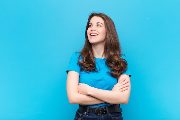 Jeune jolie femme se sentant heureuse, fière et pleine d'espoir, se demandant ou pensant, levant pour copier l'espace avec les bras croisés sur le mur bleu