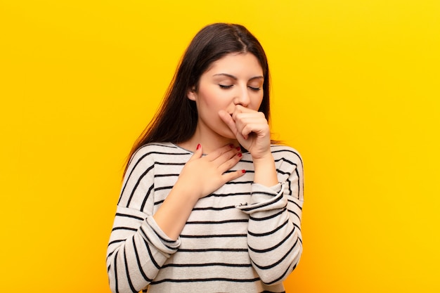 Jeune jolie femme se sentant mal avec un mal de gorge et des symptômes de la grippe, toussant avec la bouche couverte contre le mur jaune