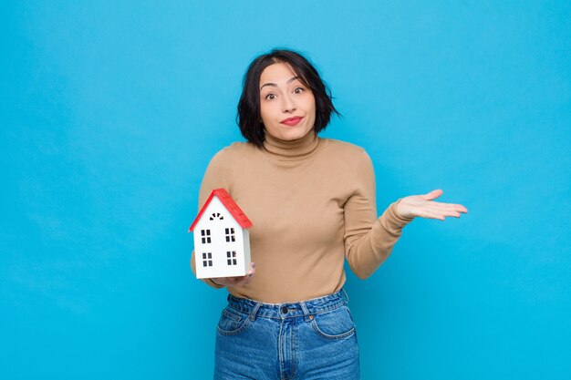 Jeune jolie femme se sentant perplexe et confuse, doutant, pondérant ou choisissant différentes options avec une drôle d'expression avec un modèle de maison