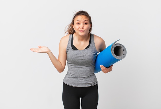 Jeune jolie femme se sentant perplexe et confuse et doutant et tenant un tapis de yoga