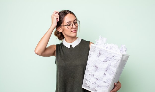 Jeune jolie femme se sentant perplexe et confuse, se grattant la tête. concept de poubelle de boules de papier