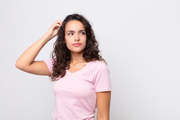 Jeune jolie femme se sentant perplexe et confuse, se grattant la tête et regardant de côté sur le mur blanc