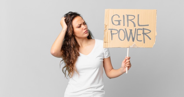 Jeune jolie femme se sentant perplexe et confuse, se grattant la tête et tenant une bannière de pouvoir des filles