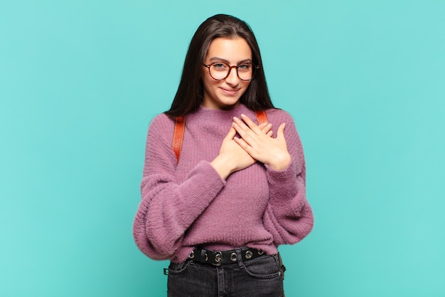 Jeune jolie femme se sentant romantique, heureuse et amoureuse, souriante joyeusement et se tenant la main près du cœur.