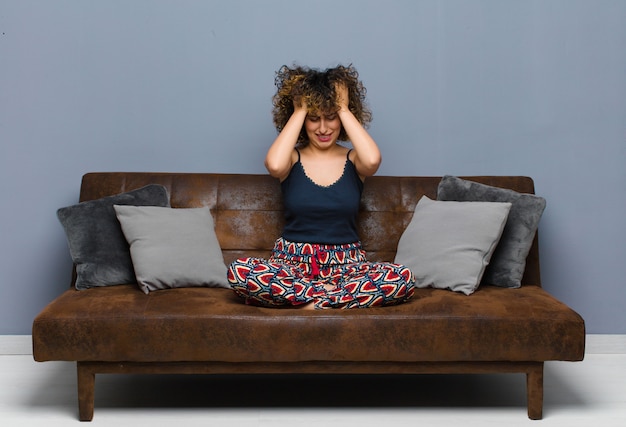 Jeune jolie femme se sentant stressée et frustrée, levant les mains à la tête, se sentant fatiguée, malheureuse et souffrant de migraine assise sur un canapé.