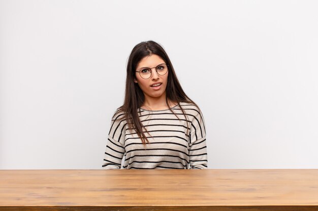 Jeune jolie femme se sentir perplexe et confus, avec une expression stupide et stupéfaite en regardant quelque chose d'inattendu assis à une table