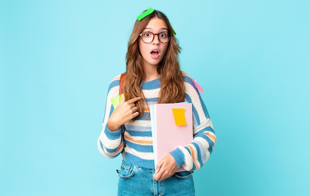 Jeune jolie femme semblant choquée et surprise avec la bouche grande ouverte, se montrant elle-même avec un sac et tenant des livres