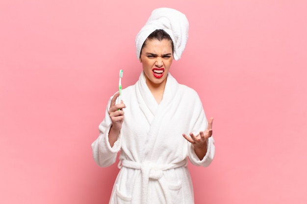 Jeune jolie femme semblant en colère, agacée et frustrée en train de crier wtf ou ce qui ne va pas avec vous. concept de peignoir et brosse à dents