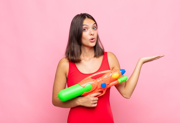 Jeune jolie femme semblant surprise et choquée, avec la mâchoire tombée tenant un objet avec une main ouverte sur le côté