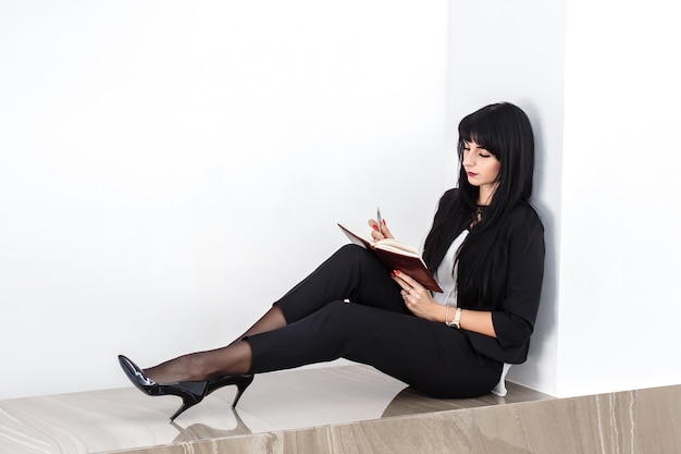 Jeune Jolie Femme Sérieuse Assise Sur Un Sol Dans Un Bureau, Lisant Dans Un Cahier.