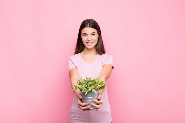 Jeune jolie femme souriant joyeusement avec un regard amical, confiant et positif, offrant et montrant un objet ou un concept avec une plante