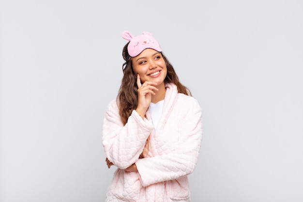 Jeune jolie femme souriant joyeusement et rêvassant ou doutant, regardant sur le côté portant un pyjama