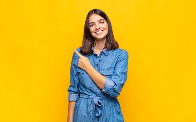 Jeune jolie femme souriant joyeusement, se sentant heureux et pointant vers le côté et vers le haut, montrant l'objet dans l'espace de copie