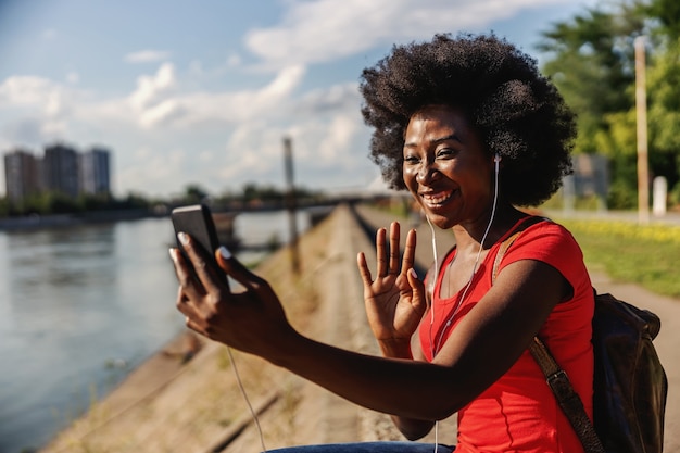 Jeune jolie femme souriante assise au bord de la rivière et ayant un appel vidéo avec un ami