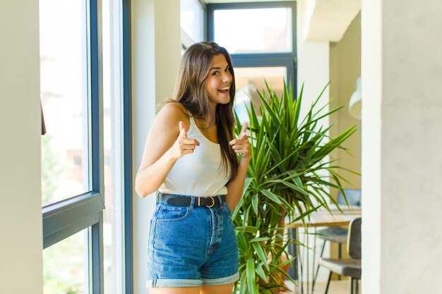 Jeune jolie femme souriante avec une attitude positive, réussie et heureuse pointant vers l'avant, faisant signe des armes à feu avec les mains