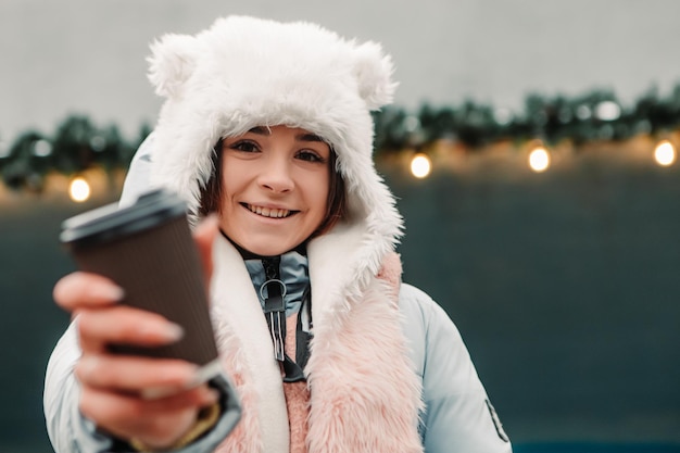 Jeune jolie femme souriante célébrant une veste chaude habillée et un bonnet de laine, buvez du thé chaud