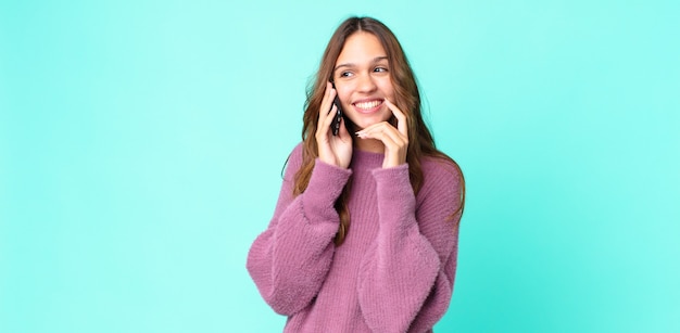 Jeune jolie femme souriante avec une expression heureuse et confiante avec la main sur le menton et à l'aide d'un smartphone