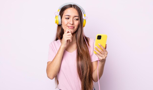 Jeune jolie femme souriante avec une expression heureuse et confiante avec la main sur le menton. casque et smartphone